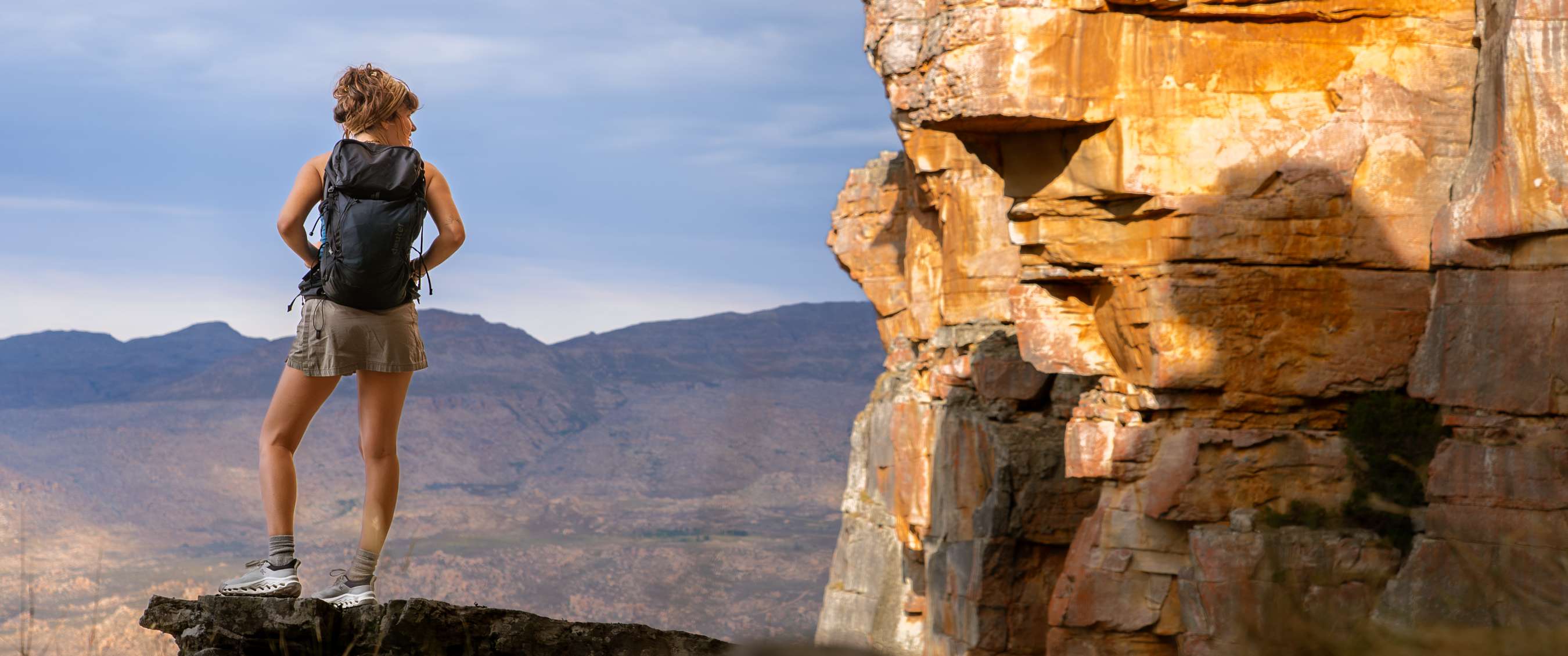 Wandelaar op uitstekende rots met bergpanorama en zonlicht op de rotswand ernaast.